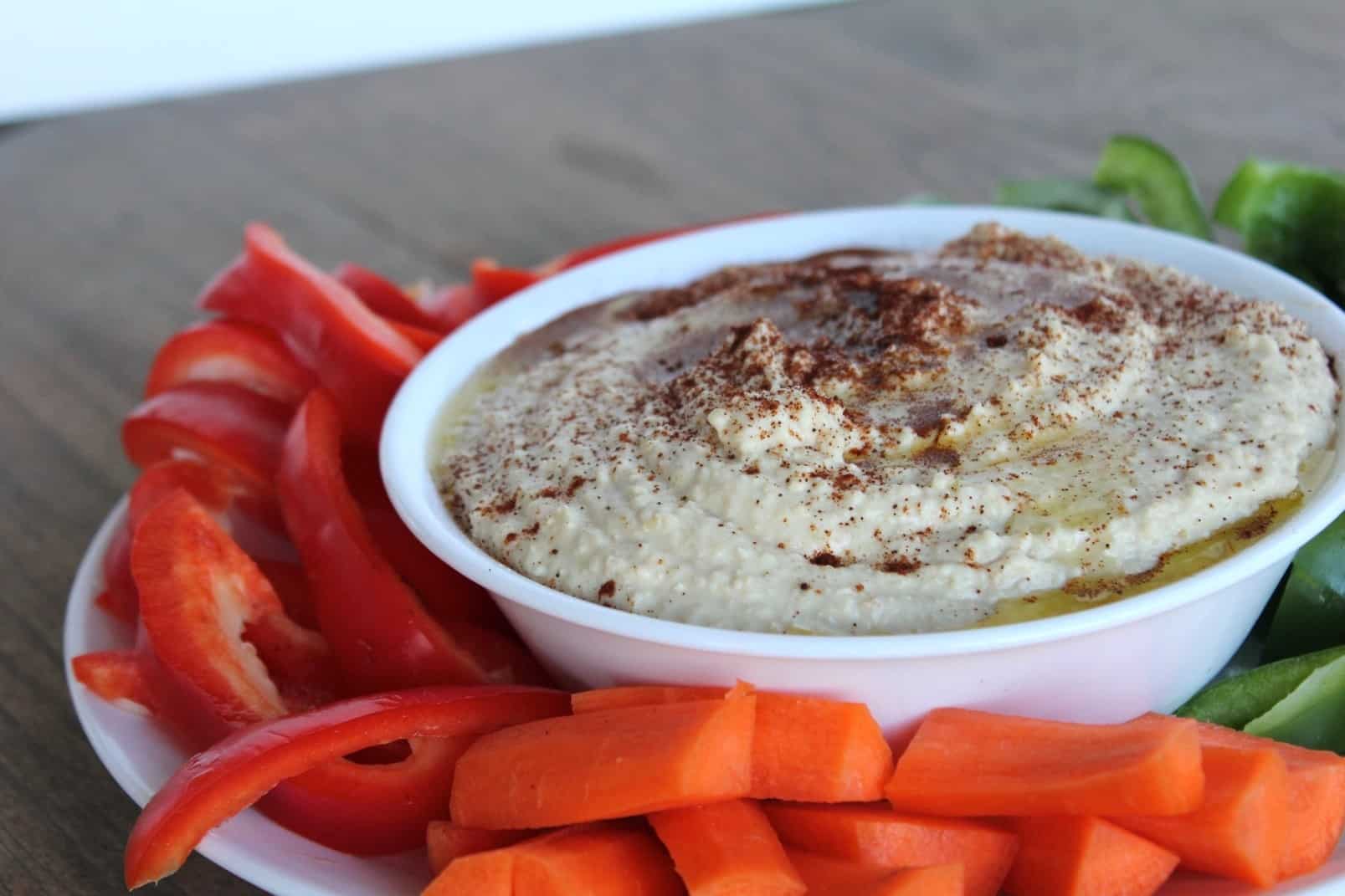 Image shows a white bowl full of homemade hummus, surrounded by carrots, red and green peppers, on a table. 