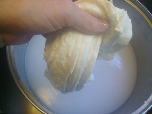 Image shows a photo of one hand holding the mozzarella cheese over the metal bowl to stretch the cheese.