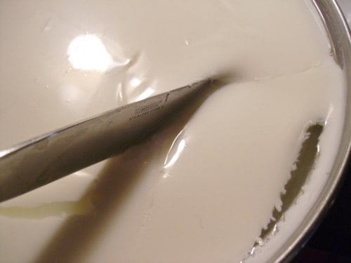 Image shows a close up, from above, of a metal spoon slicing into the milk after heating. 