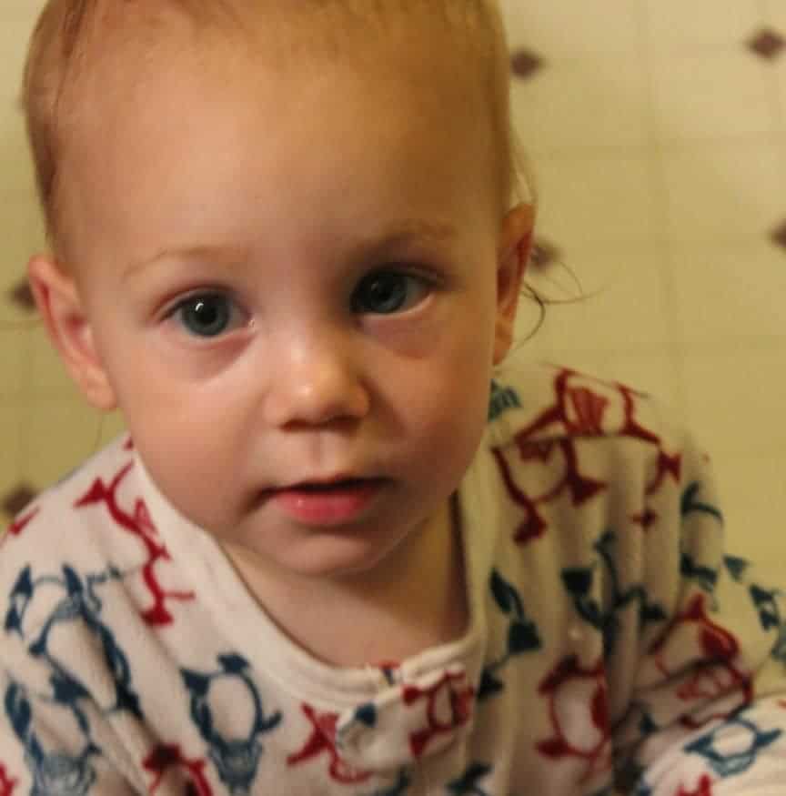 Image shows a young toddler with blue eyes looking at the camera wearing white fleece pajamas with red and blue patterns.