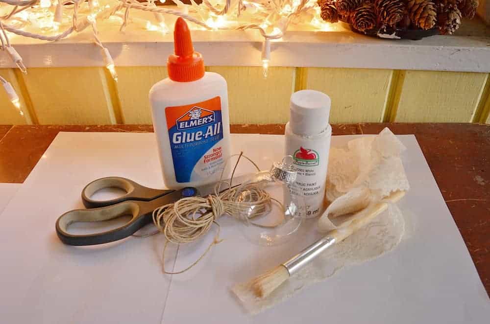 Image shows a white sheet of paper on a table, with a scissors, twine, Elmer's glue, a paint brush and lace to make a rustic lace ornament