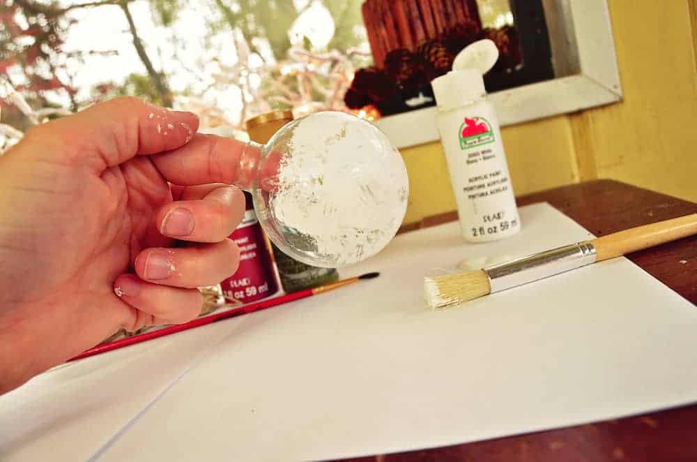 Image shows a hand holding a clear plastic ornament, partially painted with white paint. An open bottle of white paint and paintbrush sit nearby.
