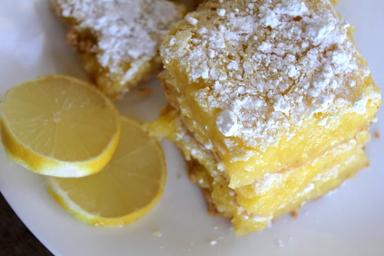 Photo, taken from above, shows a plate with several lemon bars sprinkled with powdered sugar. Two slices of lemon sit on the edge of the plate.