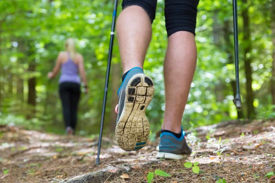 Image shows the legs of a person hiking through a wooded area. A woman hikes in front of them wearing black pants and a light blue tank top. 
