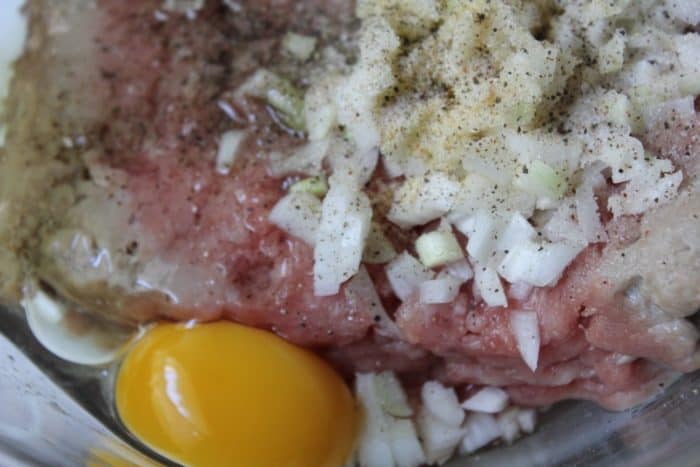 A close up image of raw ground beef, eggs, seasonings, and breadcrumbs in a bowl before being mixed into meatballs.
