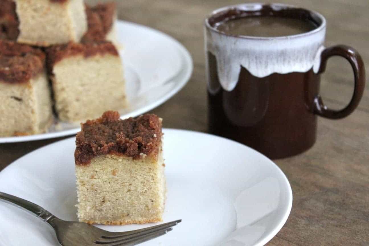 Image shows a white plate with a piece of gluten free banana coffee cake on it. Next to the plate sits a brown mug with white trim, and a plate full of more pieces of coffee cake. 