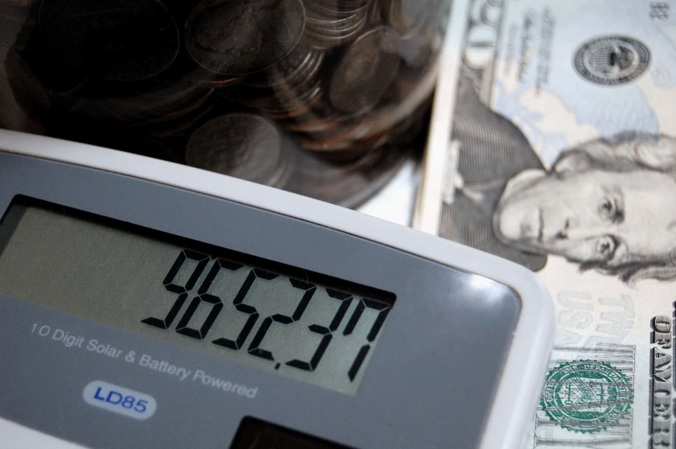 Image shows a close up of a calculator with a screen displaying numbers, with some money and a jar of coins in the background. 