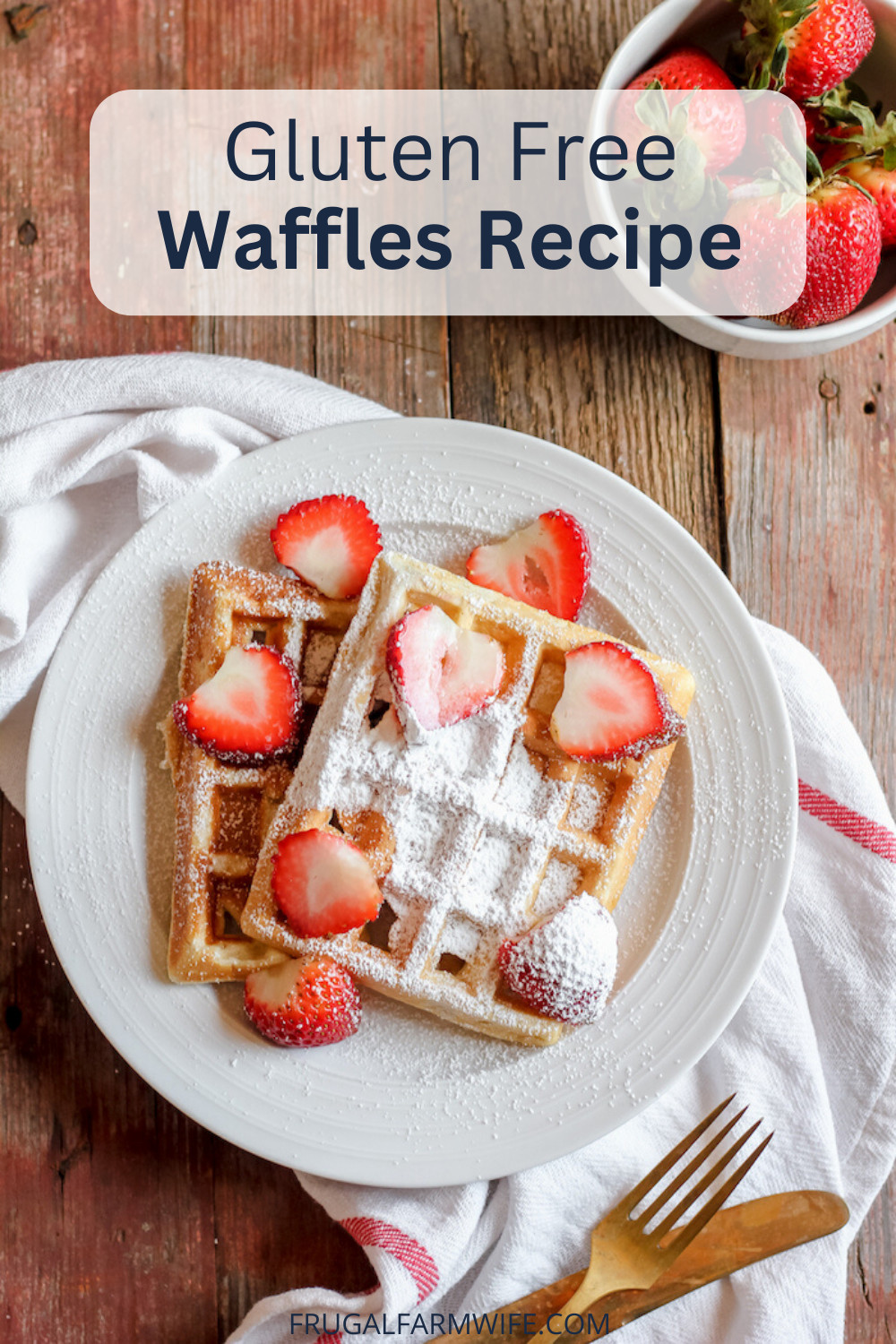 Image, taken from above, shows a plate of two gluten free waffles sprinkled with powdered sugar and covered in sliced strawberies. A bowl of more strawberries sit nearby on the table.