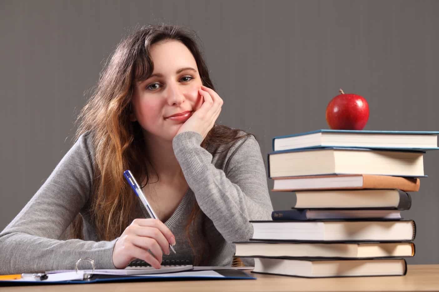Image shows a woman at a table holding a pen, sitting next to a pile of books with an apple on top. 