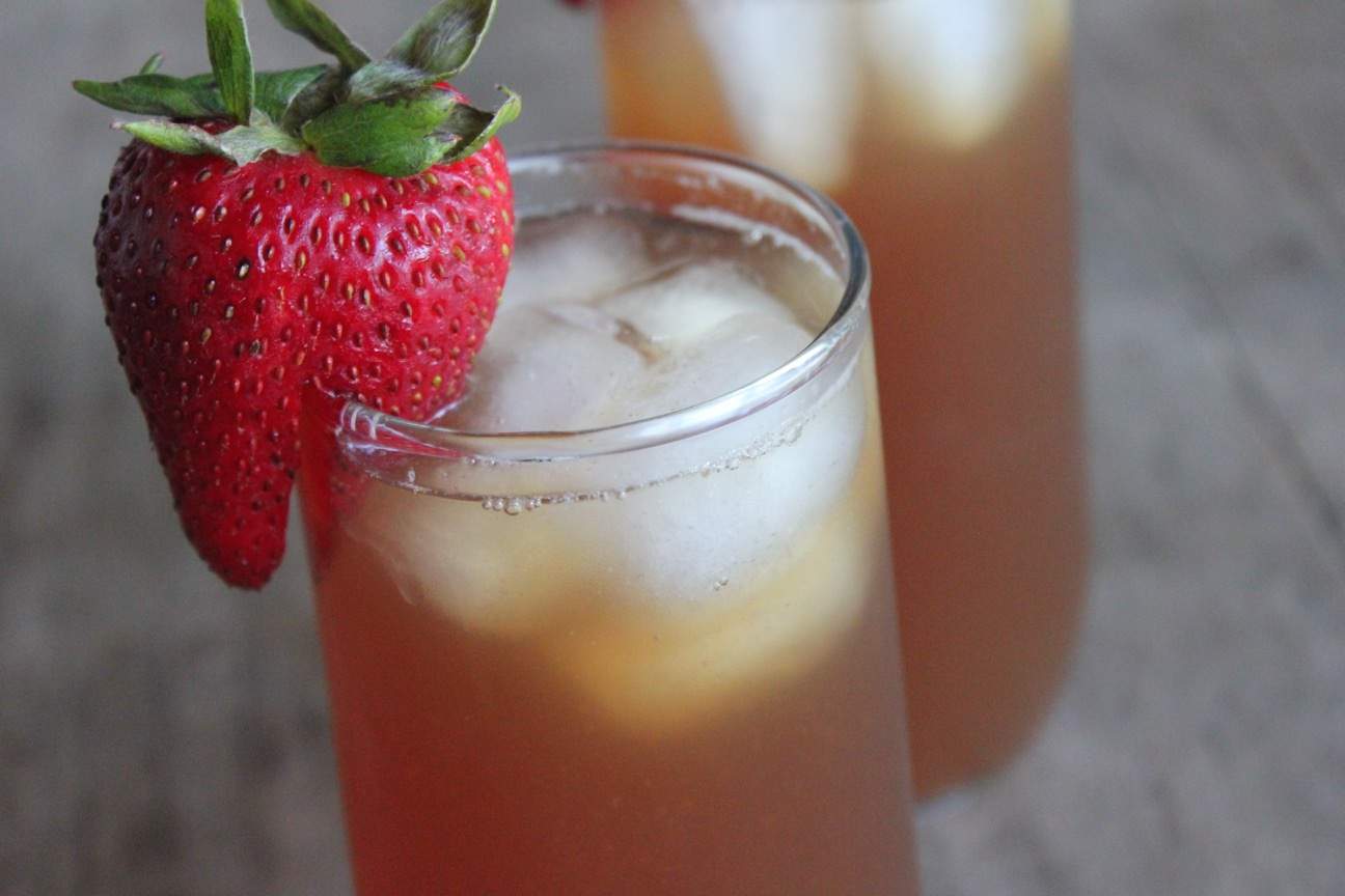 Photo shows two glasses of strawberry iced tea. The glass in front has a large strawberry sliced and resting on the rim of the glass as a garnish.