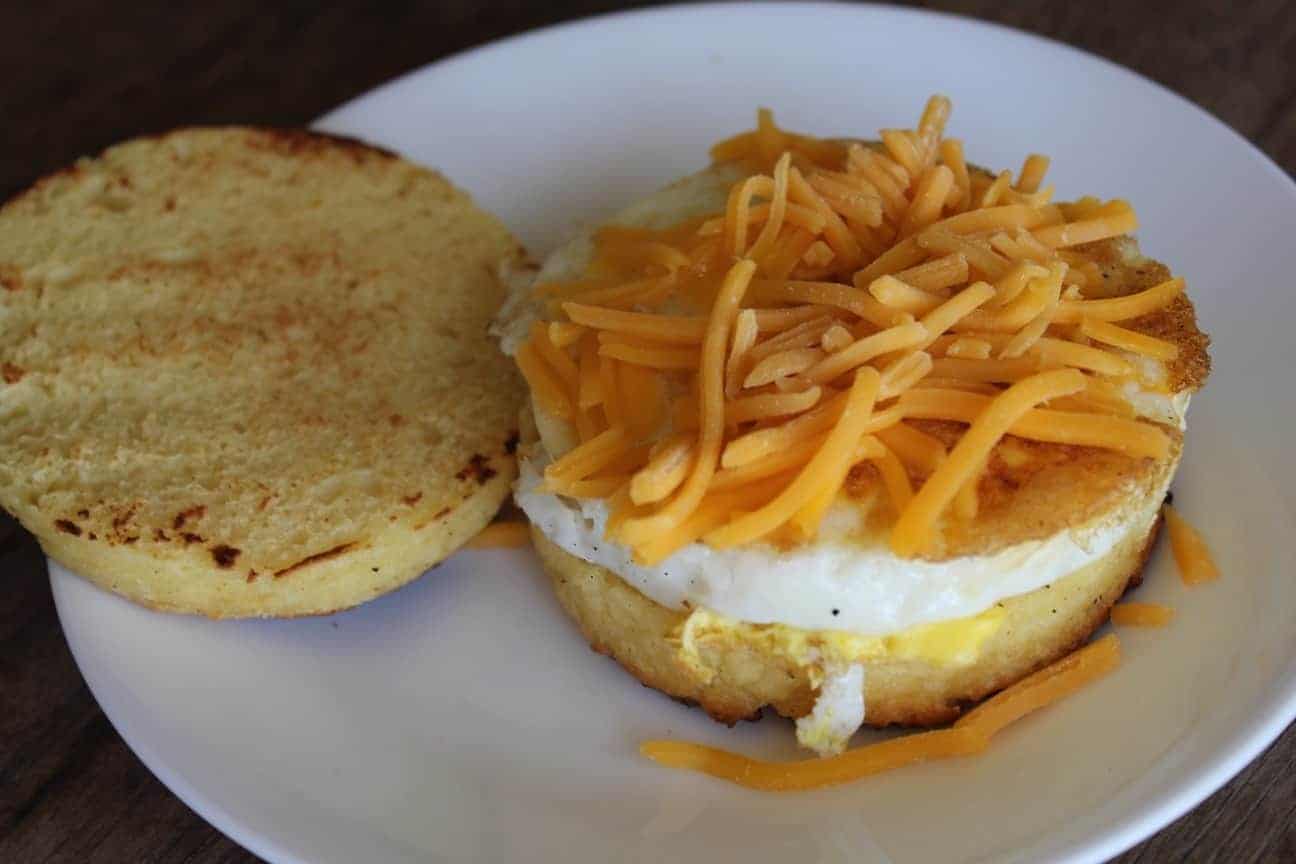 Image shows a homemade "Egg McMuffin" sandwich on a white plate with shredded cheese on it. 