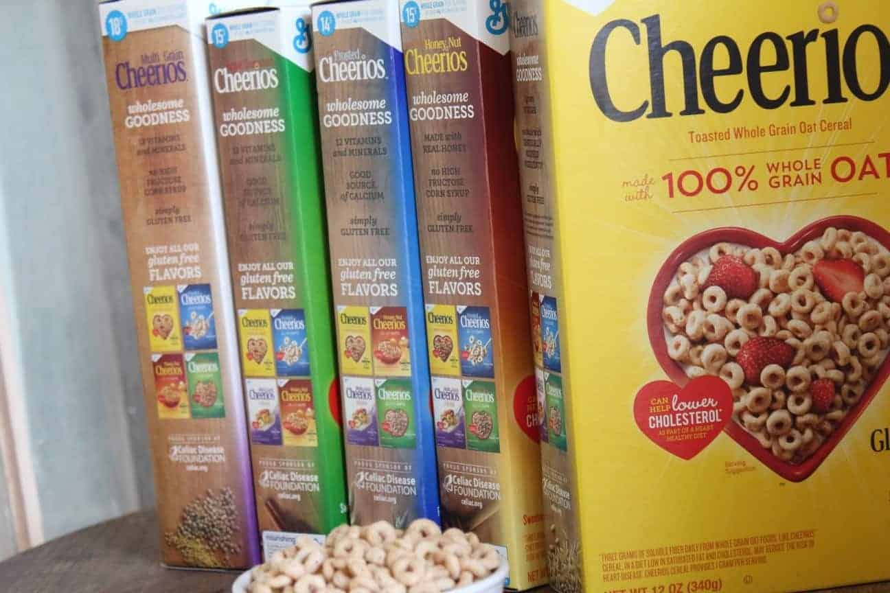 Images shows several boxes of different varieties of Cheerios on a table with a bowl of cereal sitting in front of them.