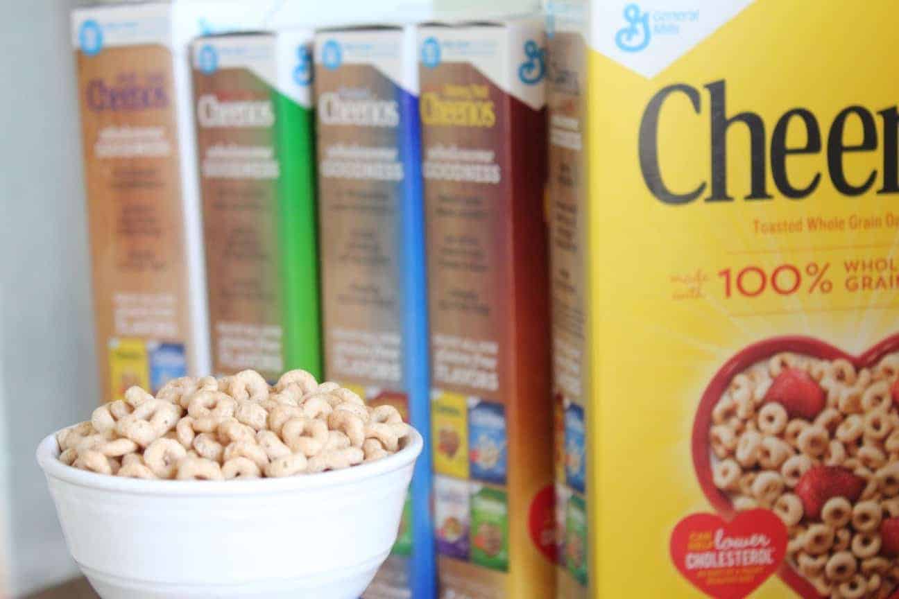 Image shows a white bowl full of Cheerios sitting on a table with several boxes of the cereal's varieties.