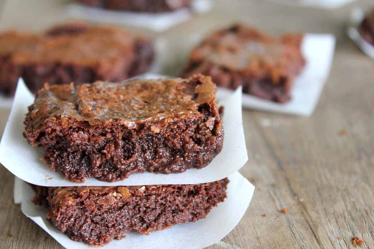 Image shows several flourless chocolate brownies on a table, each stacked on a small piece of parchment paper.