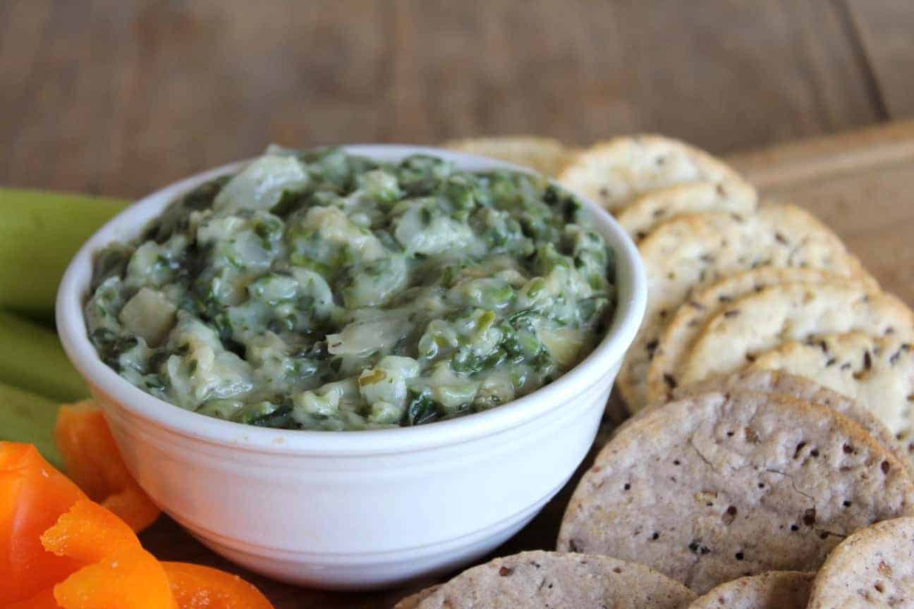 Photo shows a bowl of spinach and artichoke dip on a platter with crackers and veggies around the bowl of dip. 