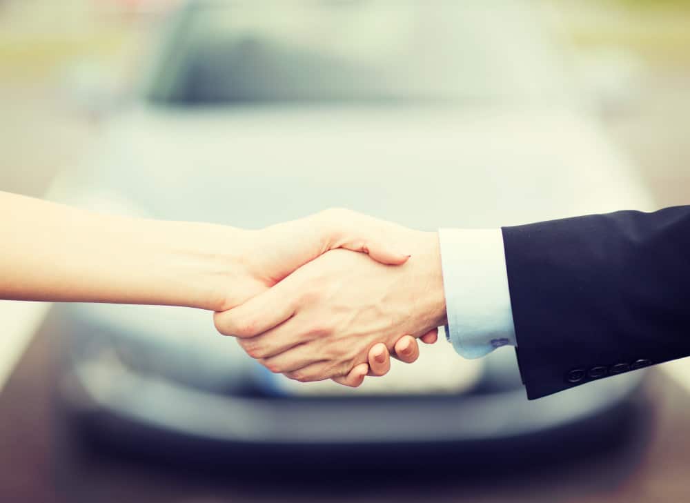 Image shows two hands clasped in a handshake in front of a blurred out grey car