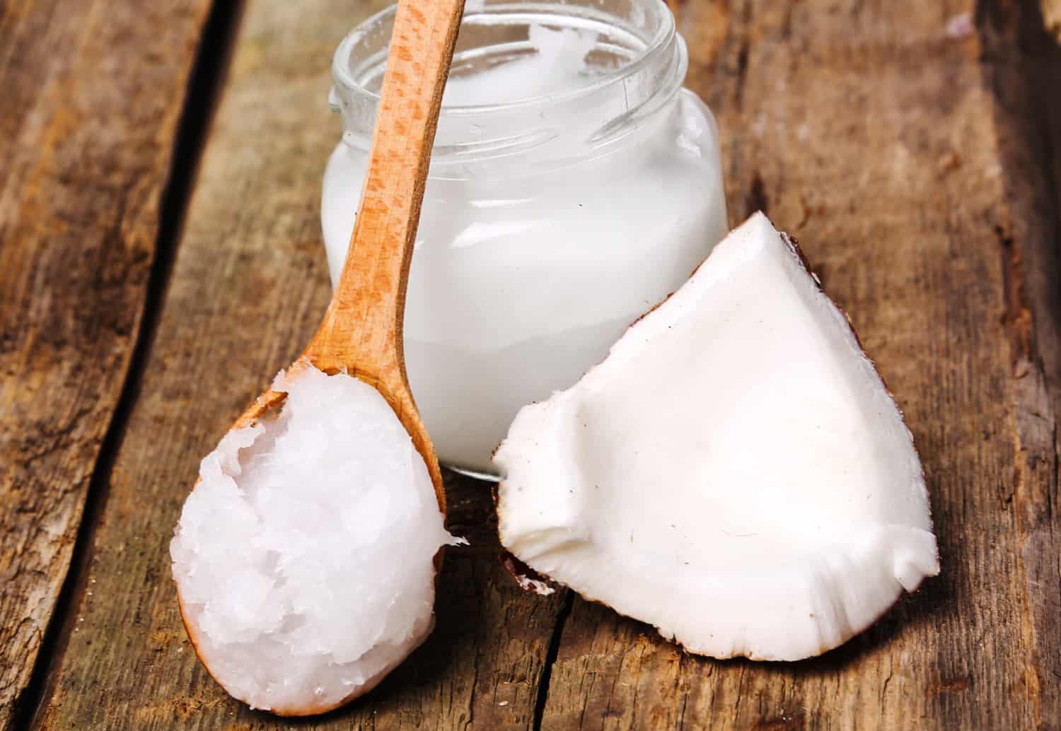 Image shows a wood table with a jar of coconut oil on it, a spoon of coconut oil and part of a coconut resting against the jar. 