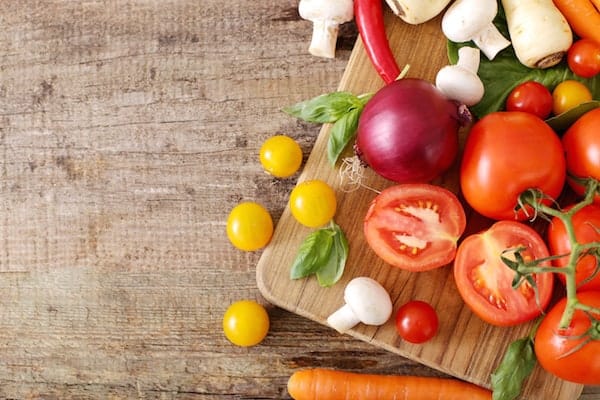 Image shows a wood cutting board on a table, with fresh veggies on it, including onion, mushroom, tomato and carrots.