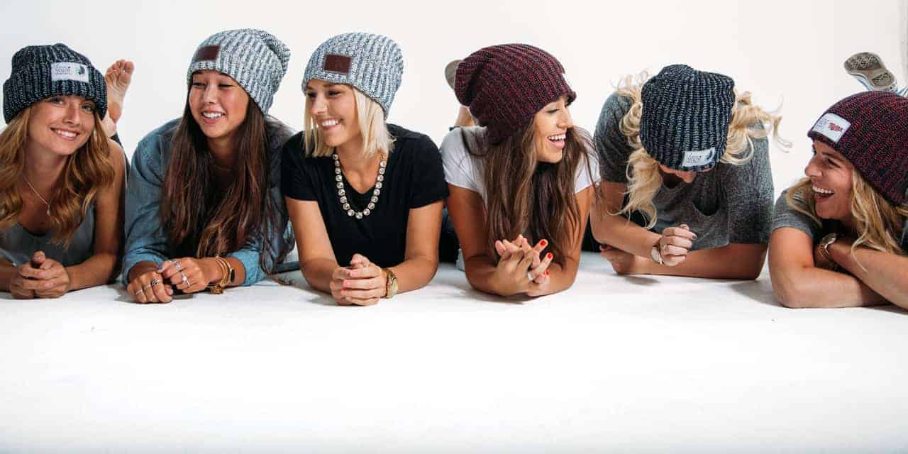 Photo shows half a dozen young women laying on their stomachs propped up on their elbows, all wearing a knit Love Your Melon hat.