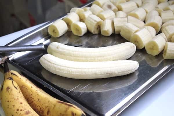 Image shows two peeled bananas sitting on a cookies sheet next to many sliced bananas.