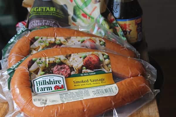 Image shows two packages of Hillshire Farm Smoked Sausage on a cutting board with vegetables and soy sauce