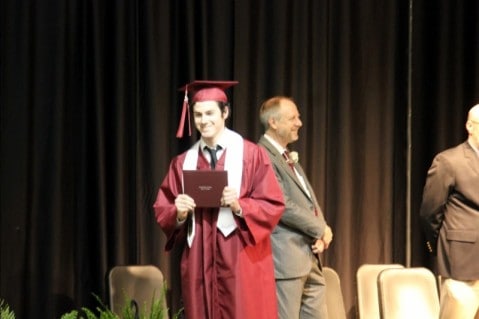 Image shows a young man in a red cap and gown holding a diploma as he crosses a stage at graduation. 