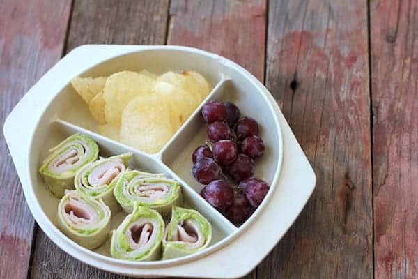 Image, taken from above, shows a divided plate sitting on a table. The plate is filled with grapes, potato chips and a rolled sandwich wrap in it.