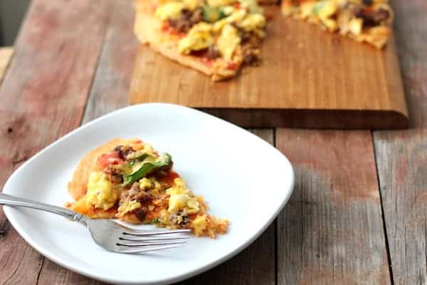Photo shows one slice of gluten-free breakfast pizza on a white plate with a fork. In the background on a piece of wood is the rest of the pizza.