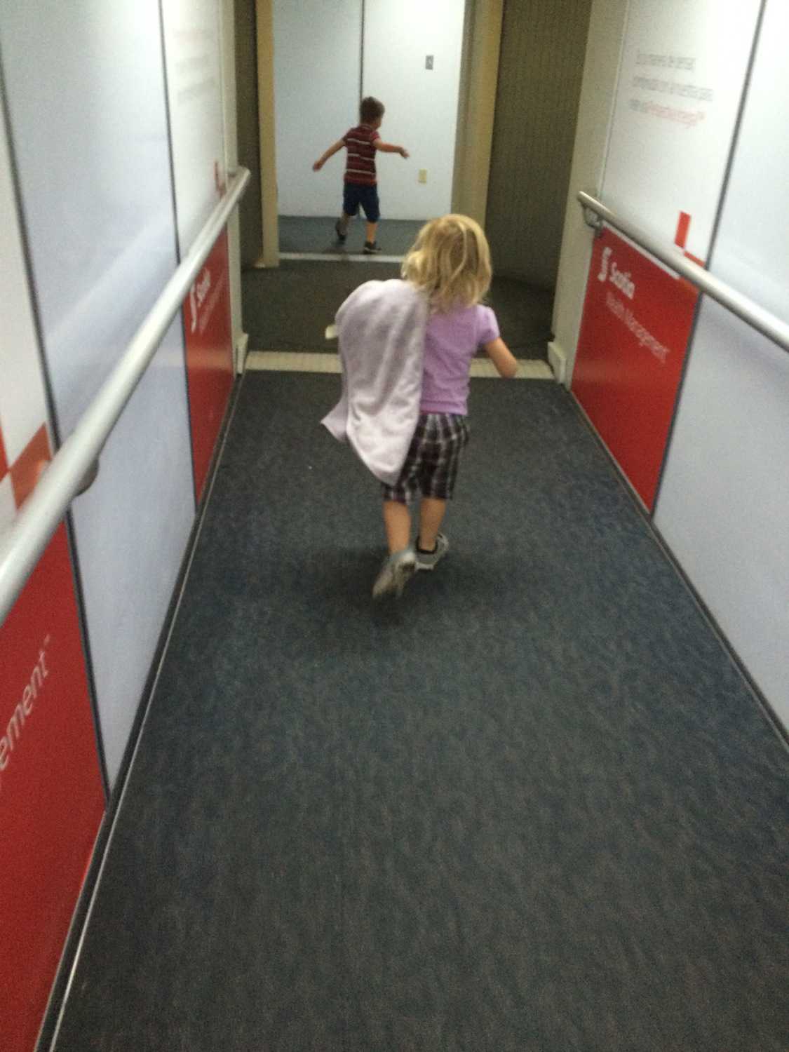 Image, taken from behind, shows two small kids running through the jetway at an airport.