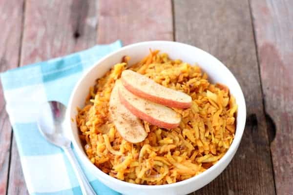 Photo shows a bowl of sweet potato hash topped with apple slices on a table next to a napkin and spoon