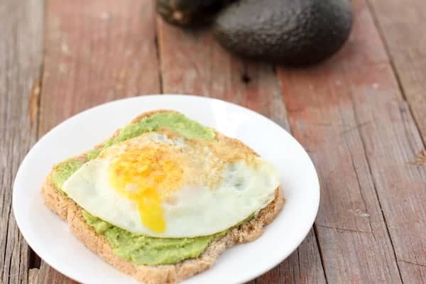 Photo shows a piece of avocado toast on a white plate with a fried egg on top. In the background 2 avocados sit on the table. 