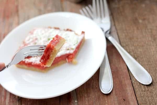 Image shows a small dessert plate with a strawberry lime bar on it. A fork hovers over the plate with a bite on it. 