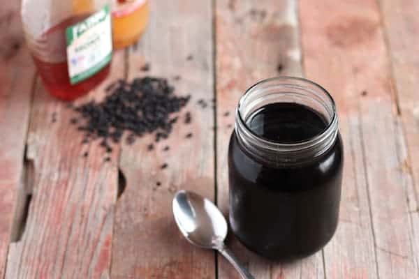 Photo shows a glass jar of elderberry syrup on a wooden table, with a spoon resting next to it. In the far corner is a bottle of honey, some dried elderberries, and vinegar.