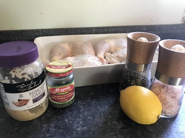 Image shows a white casserole dish on a counter with raw chicken in it. Nearby are minced garlic, capers, salt, pepper and lemon to make chicken piccata
