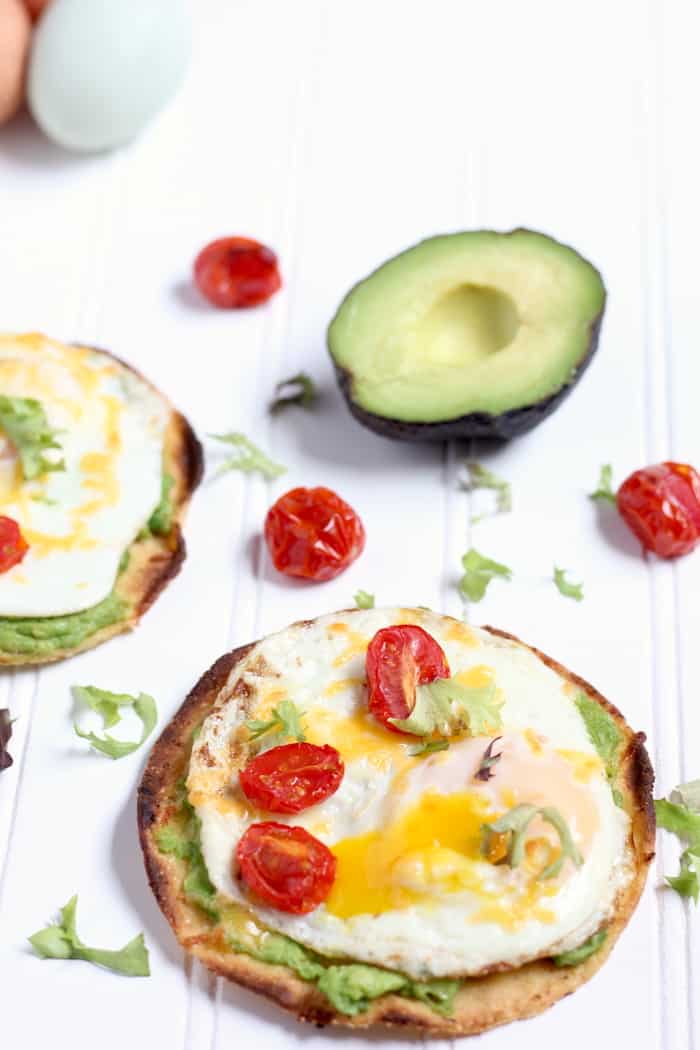 Images shows two simple breakfast tostadas on a counter- topped with egg, avocado, tomato and cheese. several tomatoes and half an avocado sit nearby.