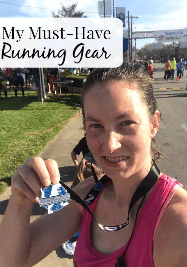 Image shows a woman standing outside along a trail, holding a running medal to the camera. Text overlay reads "My Must-Have Running Gear"
