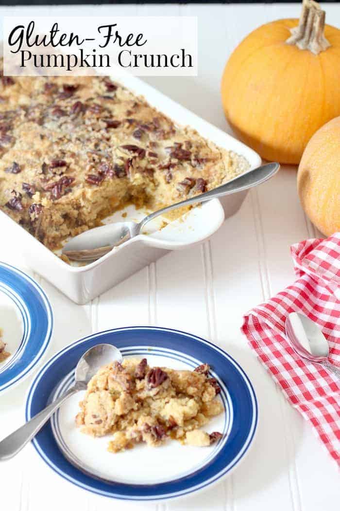 Image shows a large pan with gluten free pumpkin crunch cake on a white table. Nearby is a plate with blue trim and a serving of the pumpkin crunch. In the background are two small pumpkins. 