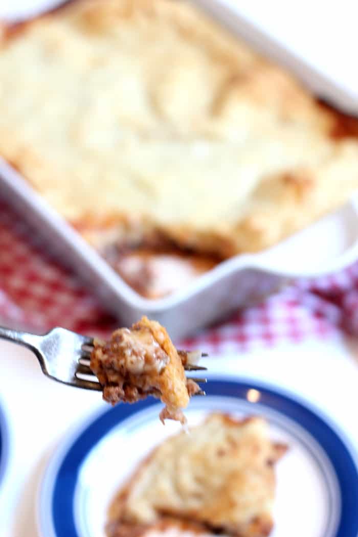 Image shows a plate with a piece of gluten free upside-down pizza on it, and a fork holding a bite. In the background is a casserole dish of more casserole.