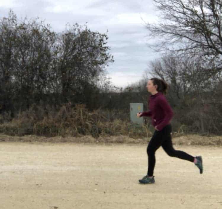 Image shows a woman running along a dirt road with headphones in her ears and a maroon jacket on.