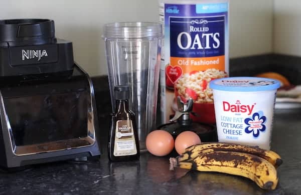 Image shows the ingredients for banana pancakes with oatmeal and cottage cheese laid out on a black countertop.