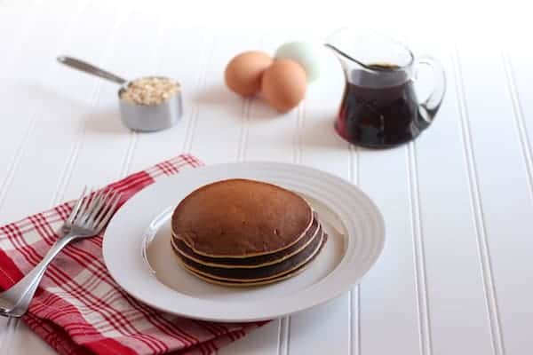 Image shows a white plate of pancakes on a white table. A red plaid cloth napkin sits nearby, along with a small pitcher of syrup, and some eggs.