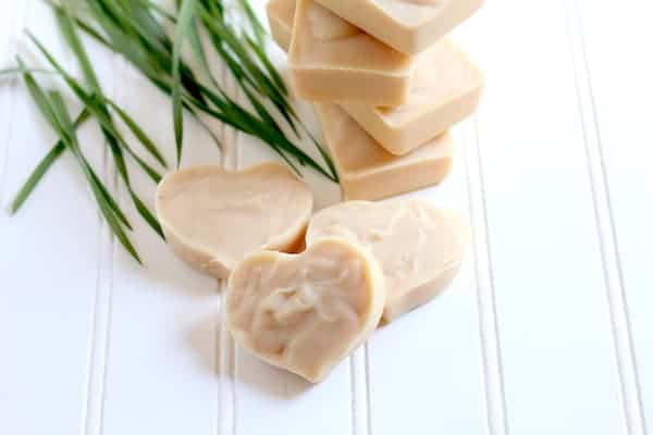 Photo shows three bars of heart shaped lemongrass soap on a white shiplap board in front of a stack of rectangle bars of soap and blades of lemongrass.