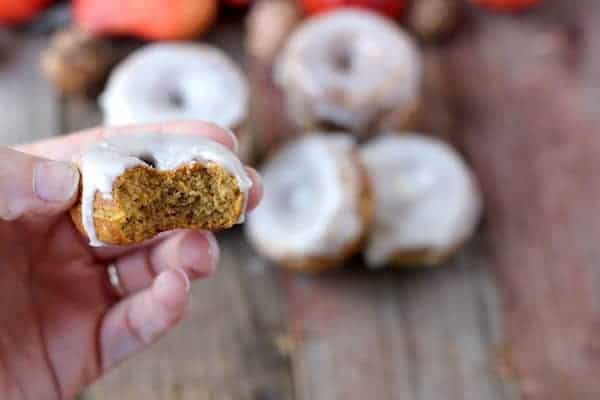 Photo shows a hand holding a pumpkin spice donut with a bite taken out of it, more blurred donuts sit in the background of the photo. 