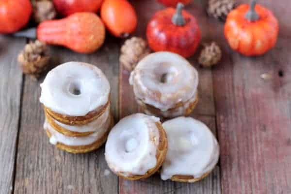 Image shows a stack of glazed pumpkin spice donuts on a table. Some small pumpkins and gourds sit in the background. 