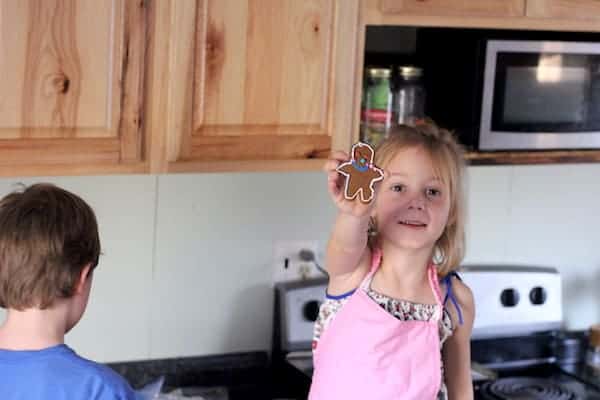 Photo shows a young girl holding up a gingerbread man cookie in a kitchen next to a young boy