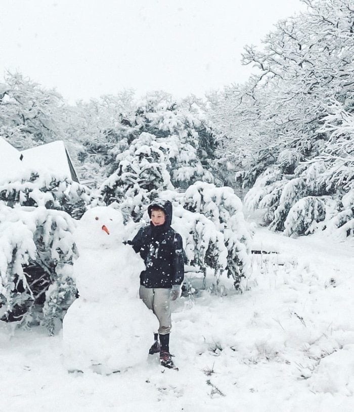 Image shows a young boy standing in the snow with a snowman