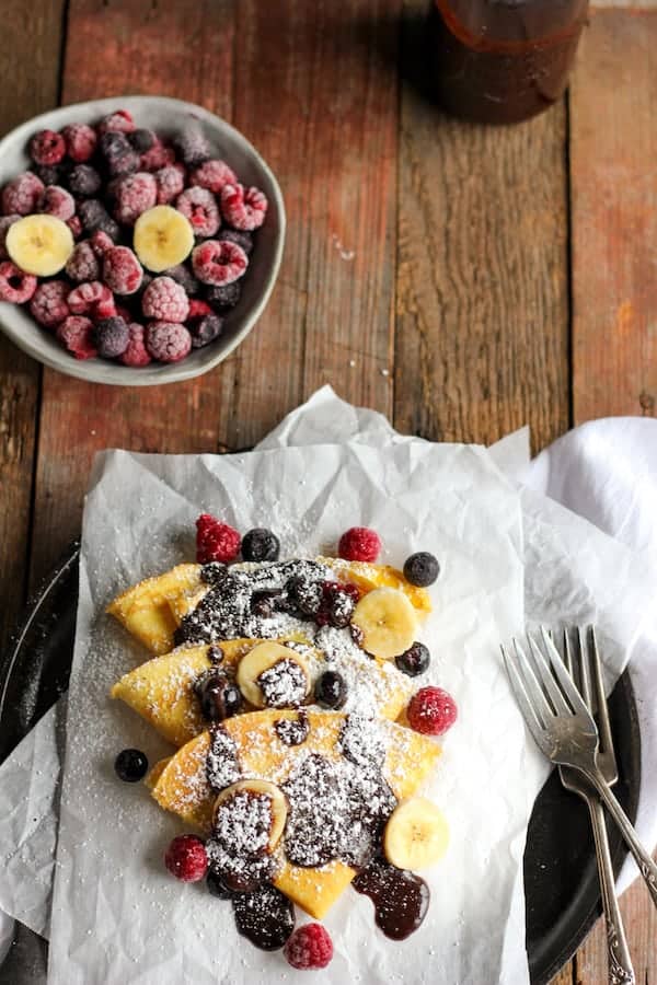 Photo shows a plate of crepes covered in chocolate and powdered sugar next to a bowl of berries