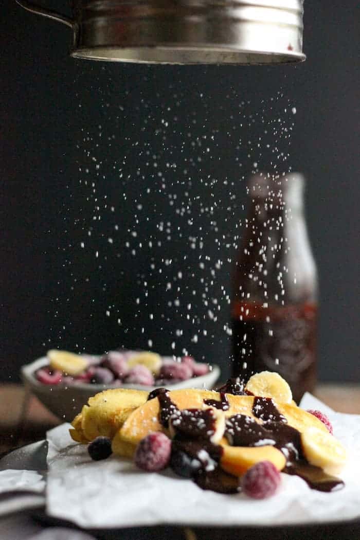 Photo shows powdered sugar being sprinkled onto a plate of crepes with chocolate and berries