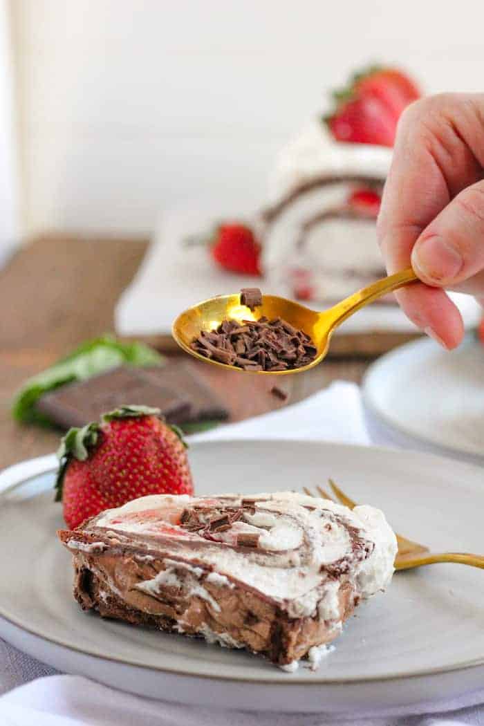 Photo shows a hand with a spoonful of chocolate pieces sprinkling them on a slice of pavlova