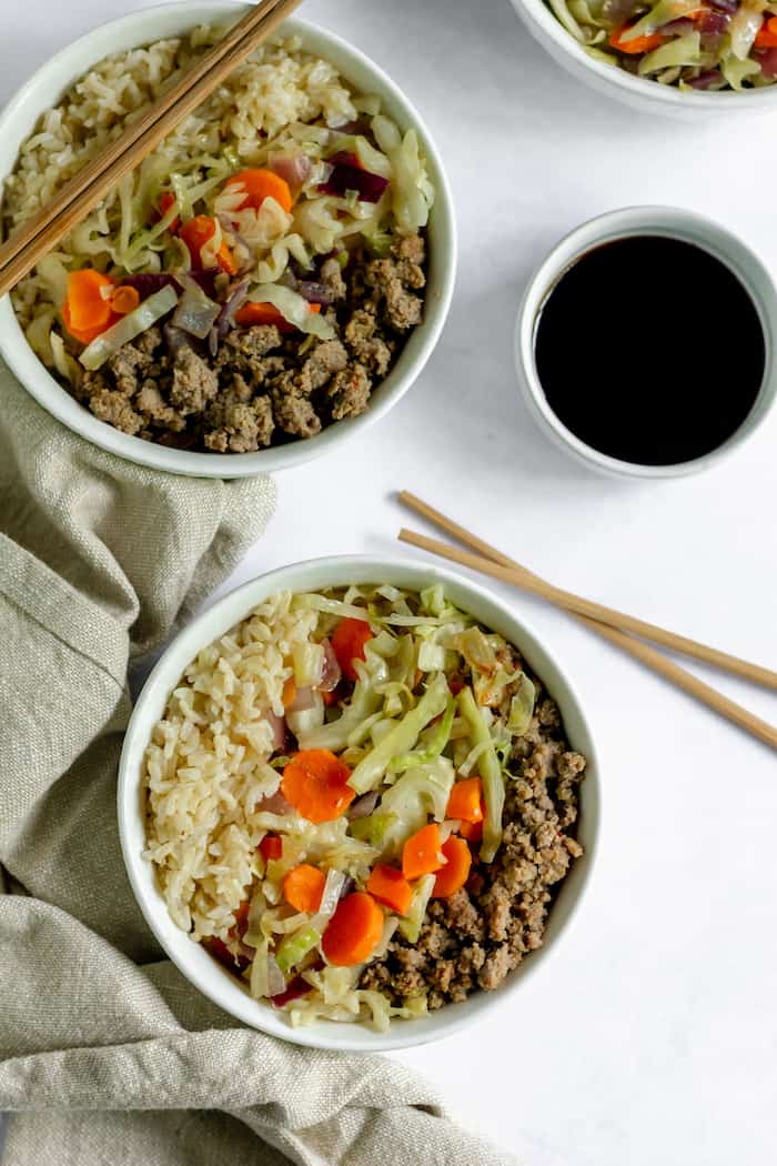 Image, taken from above, shows a close up of several bowls of egg roll in a bowl with ground meat, veggies, rice, and a small bowl of soy sauce nearby. Chop sticks rest on one bowl and on the table. 